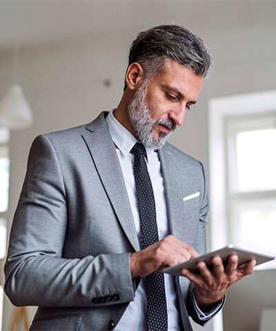 Momento Digitalizar mis gestiones- Hombre de mediana edad con barba, traje gris y corbata negra trabajado en la oficina con una tablet pensando en digitalizar las gestiones de su empresa