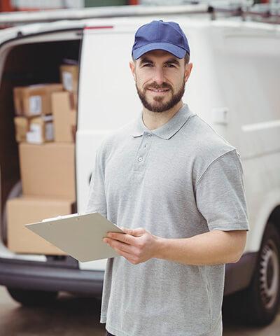 Ventajas de Seguro del Automovil para Empresas - Repartidor con gorra azul y polo gris, con una carpeta en las manos junto a una furgoneta blanca con cajas