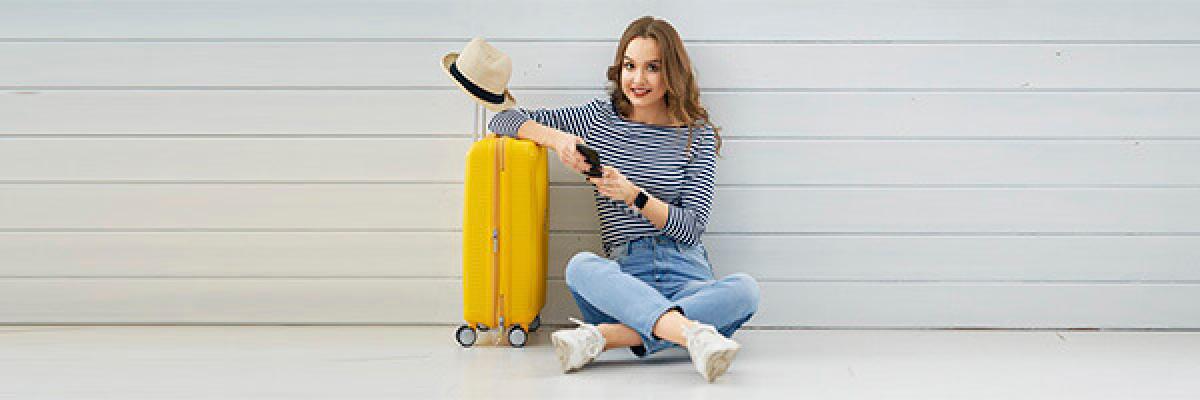 Seguro de Asistencia en Viaje - Mujer joven sentada en el suelo con el movil en la mano, junto a su maleta y un sombrero preparando un viaje Seguro de Asistencia en Viaje - Mujer joven sentada en el suelo con el movil en la mano, junto a su maleta y un sombrero preparando un viaje